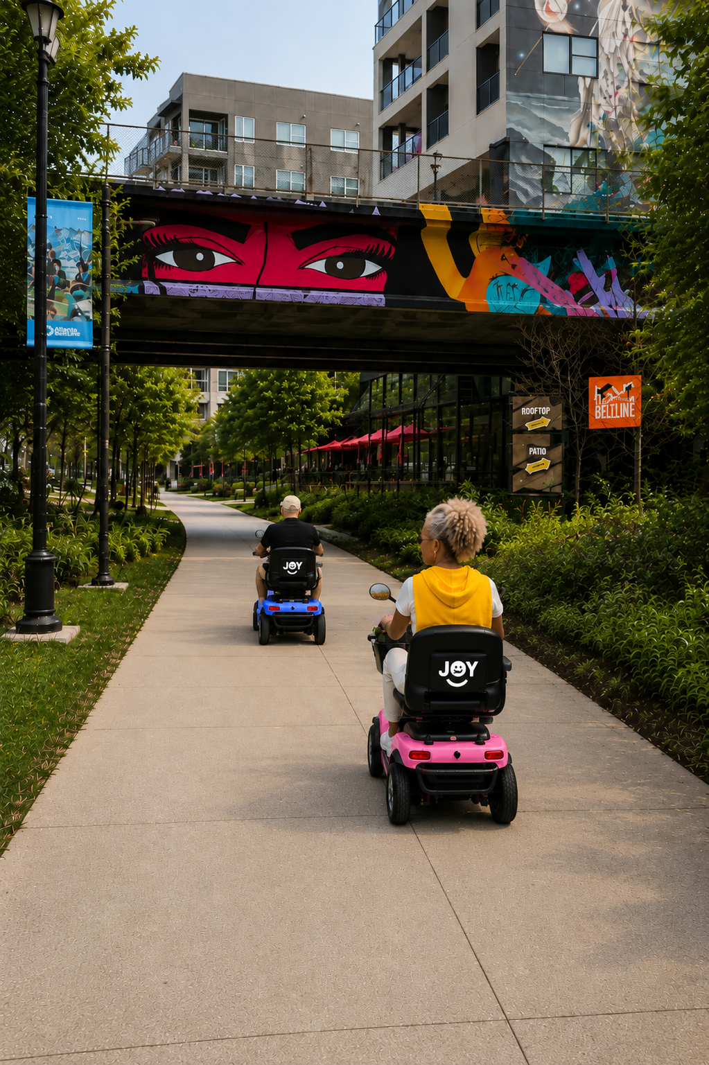 Two riders on yellow JOY mobility scooters on the BeltLine path, passing under a painted bridge.